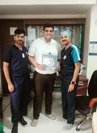 Three men standing together, two in medical scrubs, one holding a framed award or certificate.
