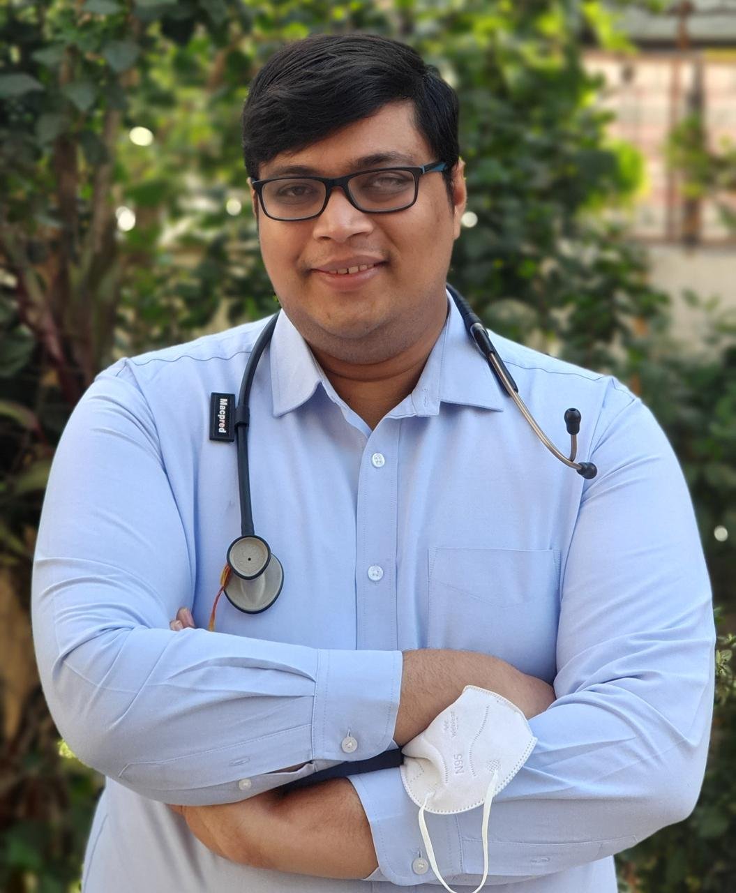 A doctor in a light blue shirt with a stethoscope around his neck, standing outdoors with a green, leafy background.