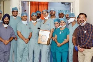A large group of medical professionals in scrubs and surgical attire, including Dr. Shirish Agrawal, pose for a photo in a hospital corridor. A woman in the center holds a large medical box.